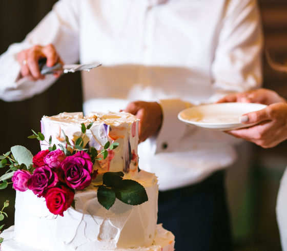 The bride and groom hold a plate and knife and cut the wedding cake with flowers and white cream. Vertical